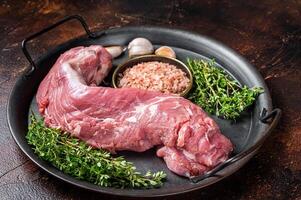 Uncooked Raw pork tenderloin fillet meat on a kitchet tray with thyme, garlic and sea salt. Dark background. Top view photo
