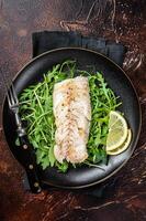Grilled cod fish fillet served with green salad in a plate. Dark background. Top view photo