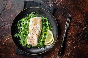 Grilled cod fish fillet served with green salad in a plate. Dark background. Top view photo