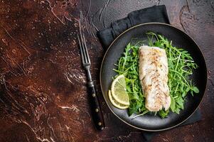 Grilled cod fish fillet served with green salad in a plate. Dark background. Top view. Copy space photo
