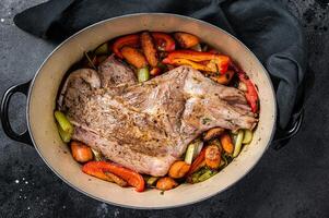Roasted lamb mutton shoulder meat in a baking dish with vegetables. Black background. Top view photo
