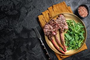 BBQ grilled lamb cutlet or mutton chop steak with arugula salad on a plate. Black background. Top view. Copy space photo