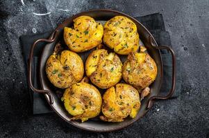 Broken potatoes baked in oil with herbs. Black background. Top view photo