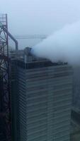 Grain processing plant premises on gray foggy day. Functioning enterprise producing heavy smoke into air. Aerial view. Vertical video