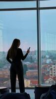 Young business woman with a phone. Rear view of a female in modern office near the large windows. Panoramic windows view with a city background. Back view. Vertical video