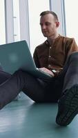 Young freelancer working on a laptop. Man in shirt and trousers sitting on a floor and using a gadget to work in office. Panoramic windows view. Vertical video