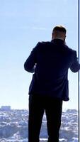 Businessman having a conversation through the phone. Young man in elegant suit standing near the large office window and talking the phone. Rear view. Vertical video