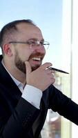 Smiling business worker talking to someone in office. Portrait of entrepreneur in glasses and costume sitting at his desk on the background of window in the city. Vertical video