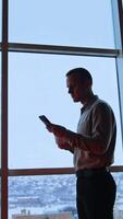 Calm serious man stands at the panoramic window holding a cup and a phone. Male scrolling smartphone screen looking for information. Vertical video