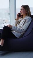 Resilient positive lady sits in bean bag chair talking on the phone. Woman waves her hand with glasses during conversation. Panoramic windows in blur at backdrop. Vertical video