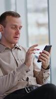 chat conversation at smartphone. Middle-aged man talks and smiles and waves his hand sitting in chair near window. Blurred backdrop. Vertical video