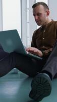 Young man sitting on floor near the window with a laptop. Happy man winning something while using a wireless computer. Vertical video