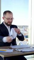 Nervous businessman in office. Irritated man entrepreneur sitting at table and tearing papers throwing them to the sides on the background of window. Vertical video