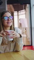 Relaxed lady drinks tea enjoying lunch time rest. Young smiling woman sitting at the table and looking in the window. Vertical video