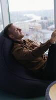 Young businessman relaxing near the window. Handsome man sitting in a cosy chair and having a conversation with the city window view. Profile view. Vertical video