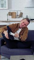Young man rests on a couch at home. Funny man brushing his socks while sitting on a sofa next to laptop at home. Quarantine concept. Vertical video