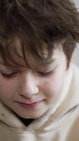 Dark-haired teenage boy looking down and then straight into camera. Close up. Portrait of a kid smiling. Light backdrop. Vertical video