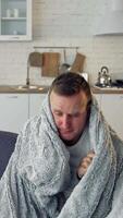 Man suffering from flu. Young sick man sitting on a sofa warming with blanket in the kitchen. Miserable man having a headache. Covid-19. Isolation at home. Vertical video