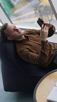 Man rests in a soft chair with a phone. Young man sitting near the window with a city transport background and using his mobile phone. Cup of coffee on small table. Top view. Vertical video