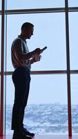 Young man standing tall at panoramic window with a cup and phone in hands. Full-length portrait of a male worker in office during lunch time. Vertical video