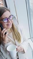 Smiling young lady stands at the panoramic window with a cup in her hand. Woman speaks on the phone and drinks tea. High angle view. Vertical video