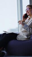 Young business lady sits in chair and talks on the phone. Left-handed woman making notes in her paper notebook. Panoramic windows in blur at backdrop. Vertical video
