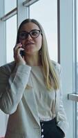 Positive young woman talking on the phone and smiling. Lady in glasses stands at the big window having phone conversation. Vertical video