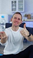 Man with a bottle of water at home. Young man in white t-shirt sitting on a floor and showing a bottle after doing fitness exercises. Vertical video