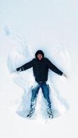 Adult man making snow angels. High angle view of happy man lying on snow and creating snow angel figure. Vertical video