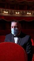Visitor in suit and medical mask sitting in comfortable chairs alone on dark theater background. Male among rows of empty red chairs. Pandemic concept. Vertical video