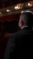 Theater actor speaks on a stage in medical mask. Announcer in suit stands in front of empty theater hall. Pandemic concept. Vertical video