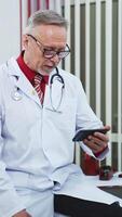 Happy satisfied mature doctor is working with patient via phone. Looking at phone in modern clinic office. Closeup. Vertical video