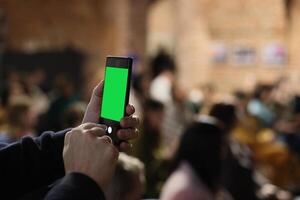 A person is holding a smartphone that features a green screen, surrounded by a vibrant crowd scene photo