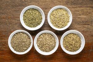 herb spices - dried leaves of parsley, rosemary, thyme, oregano and basil, collection of small ceramic bowls on a rustic wood, top view photo