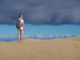 A man traveler tourist wearing sunglasses and backpack walks through the desert at the background of cloudy sky. Dune in Maspalomas, Gran Canaria, Spain photo