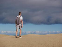 A man traveler tourist with a backpack walks through the desert at the background of cloudy sky. Dune in Maspalomas, Gran Canaria, Spain photo