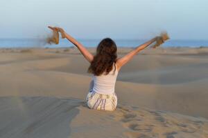 Back view of a girl seating on a sand and scattering sand from raised hands at the background of a sea at a golden hour before sunset. Dune in Maspalomas, Gran Canaria. Touristic travel concept photo