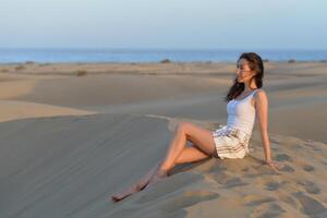 A girl seating on a sand at the background of a sea at a golden hour before sunset. Dune in Maspalomas, Gran Canaria. Touristic travel concept photo