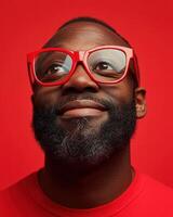 A man with a beard smiles confidently while wearing large red glasses in front of a bright red background photo