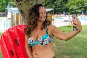 beautiful smiling middle-aged woman taking a selfie in swimsuit sitting on an inflatable under a tree photo