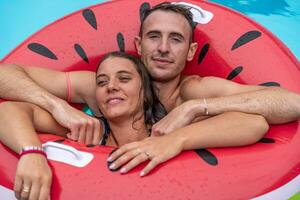 young couple in swimsuits having fun in the splashing blue water of a swimming pool inside a red inflatable life ring photo