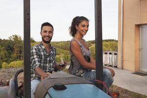 young couple of winemakers toasting with two glasses of wine sitting on an old crawler tractor at sunset photo
