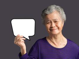 An elderly Asian woman is holding a blank speech bubble with a smile and looking at the camera while standing on a gray background photo