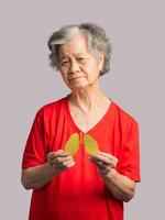A senior woman holding a lungs shape made from yellow paper while standing on a red background photo