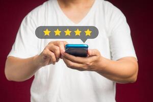 Satisfaction and feedback surveys. A man in a white casual using a smartphone gives the five-star icon a rating of very impressed for service while standing over a red background photo