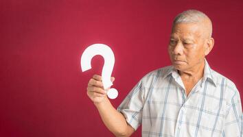 Portrait of a senior man holding and looking at a white question mark while standing with a red background photo