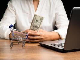 Close-up of hands holding US banknotes with a mini shopping trolley, and laptop on a wooden table while sitting in the office photo