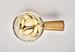 Pile of pistachio nuts in small glassware pot cup with wooden handle isolated on horizontal white background from top angle view. photo