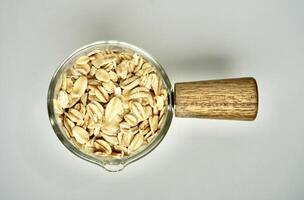 Pile of organic rolled oats food ingredients in small glass pot cup with wooden handle isolated on horizontal white background from top angle view. photo