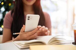 A close-up of an Asian woman using her smartphone while doing homework at a table in a cafe. photo